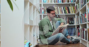 Caucasian student man reading book, sitting among stack of books on library floor, side view.