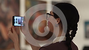 Caucasian student woman wearing eyeglasses take photo with smartphone in art gallery. Visiting excursion in museum