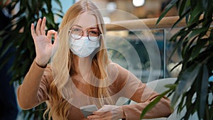 Caucasian successful business woman girl in medical mask indoors with mobile phone success online shopping showing ok