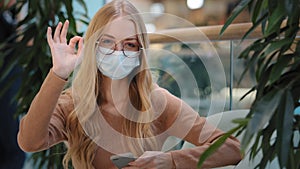 Caucasian successful business woman girl in medical mask indoors with mobile phone success online shopping showing ok