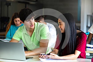 Caucasian teen boy and Asian teenage female high school students in class talking and working together using laptop.