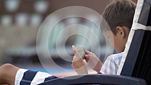 Caucasian teen boy lying on lounger operating mobile phone at swimming pool