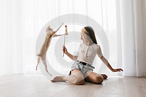 Caucasian teen girl sitting on the floor playing with a cat cornish rex with a stick teaser in the house