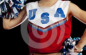 A caucasian teen girl wearing sleeveless shirt with USA printed on top against dark background is holding two pom poms.