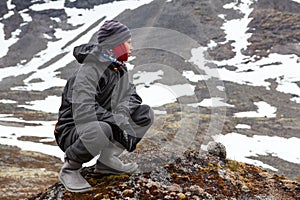 Caucasian teenage boy dressing warm hiking outfit for mountaineering, young man sitting on big stone and looking far away in