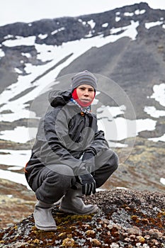 Caucasian teenage boy dressing windproof and waterproof hiking outfit for mountaineering, young man sitting on rock and looking at