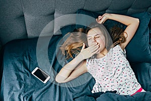 A Caucasian teenage girl in pajamas has just woken up, lies in bed next to the phone and yawns with her hand over her mouth and do