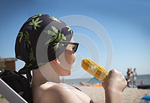 A Caucasian teenager boy in a bandana and sun glasses eats boiled corn on the beach . The concept of relaxation and tranquility.
