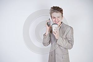 Caucasian teenager in a gray business suit stands with a mobile phone and a paper Cup of coffee on a white background in the Studi