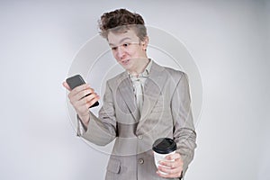 Caucasian teenager in a gray business suit stands with a mobile phone and a paper Cup of coffee on a white background in the Studi