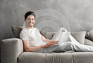 Caucasian unshaved man 30s in casual clothing using notebook, while lying on cozy sofa in gray apartment