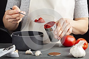 A caucasian woman is adding tomato paste from a can into a dish she is preparing in a nonstick pot.