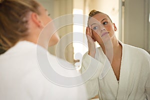 Caucasian woman in bathroom wearing bathrobe, looking in mirror and moisturising face