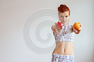 Caucasian woman in black and white sportswear holding apple and orange with undesirable face on white wall background