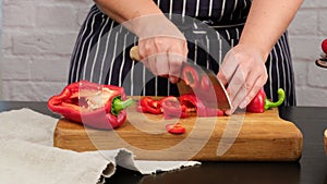 Caucasian woman in blue apron cuts ripe red bell pepper into pieces on wooden cutting board