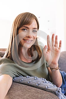 Caucasian woman with brown hair waves at the camera on video call, sitting comfortably at home