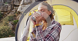 Caucasian woman camping, sitting outside tent drinking coffee and smiling in rural mountain setting