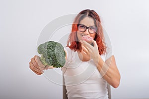 Caucasian woman chooses between vegetables and fast food. Redhead girl eating pochik and holding broccoli on a white