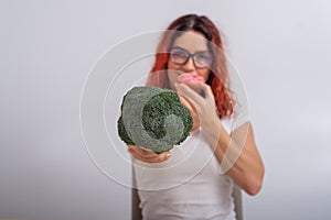 Caucasian woman chooses between vegetables and fast food. Redhead girl eating pochik and holding broccoli on a white