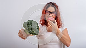 Caucasian woman chooses between vegetables and fast food. Redhead girl eating pochik and holding broccoli on a white