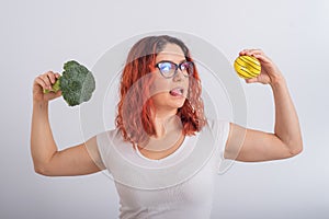 Caucasian woman chooses between vegetables and fast food. Redhead girl holding broccoli and donut on a white background.