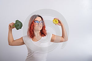 Caucasian woman chooses between vegetables and fast food. Redhead girl holding broccoli and donut on a white background.