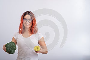 Caucasian woman chooses between vegetables and fast food. Redhead girl holding broccoli and donut on a white background.