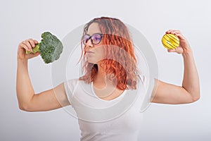 Caucasian woman chooses between vegetables and fast food. Redhead girl holding broccoli and donut on a white background.