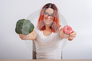 Caucasian woman chooses between vegetables and fast food. Redhead girl holding broccoli and donut on a white background.