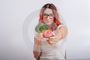 Caucasian woman chooses between vegetables and fast food. Redhead girl holding broccoli and donut on a white background.