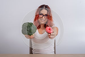 Caucasian woman chooses between vegetables and fast food. Redhead girl holding broccoli and donut on a white background.