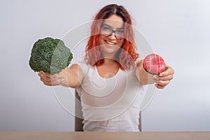 Caucasian woman chooses between vegetables and fast food. Redhead girl holding broccoli and donut on a white background.