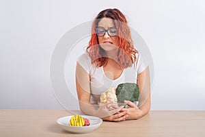 Caucasian woman on a diet dreaming of fast food. Redhead girl chooses between broccoli and donuts on white background.