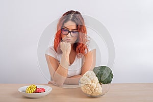 Caucasian woman on a diet dreaming of fast food. Redhead girl chooses between broccoli and donuts on white background.