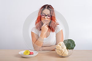 Caucasian woman on a diet dreaming of fast food. Redhead girl chooses between broccoli and donuts on white background.