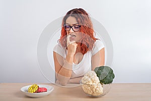 Caucasian woman on a diet dreaming of fast food. Redhead girl chooses between broccoli and donuts on white background.