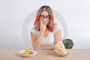 Caucasian woman on a diet dreaming of fast food. Redhead girl chooses between broccoli and donuts on white background.
