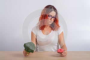 Caucasian woman on a diet dreaming of fast food. Redhead girl chooses between broccoli and donuts on white background.