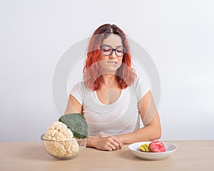 Caucasian woman on a diet dreaming of fast food. Redhead girl chooses between broccoli and donuts on white background.