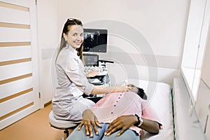 Caucasian woman doctor examining thyroid gland of young African woman using ultrasound scanner machine. Doctor runs