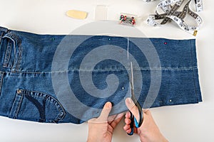 Caucasian woman hands cutting out a folded in half blue denim capris with scissors on a white table. Shorten the jeans with