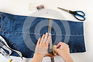 Caucasian woman hands drawing a cut line on a folded in half blue denim capris on a white table. Shorten the jeans with scissors
