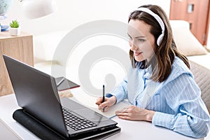 Caucasian woman with headphones writes in notebook, looks at laptop screen, remote work at home, home training