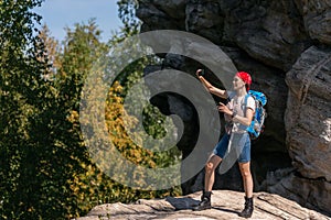 Caucasian woman hiker with backpack and red bandana does selfie standing on a rock in sunny day in summer, full length