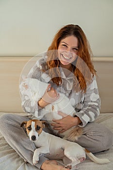 Caucasian woman holding a white fluffy cat and Jack Russell Terrier dog while sitting on the bed. The red-haired girl
