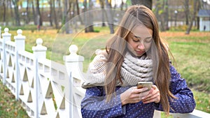 Caucasian woman with knitten scarf using smart phone, typing something during walking in autumn park. video 4k