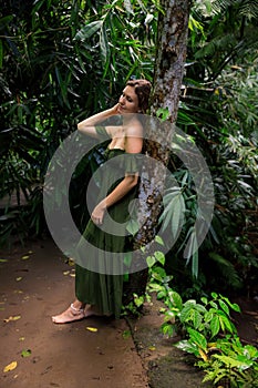 Caucasian woman leaning against a tree in tropical rain forest. Beautiful woman wearing long green dress. Walking trail in jungle