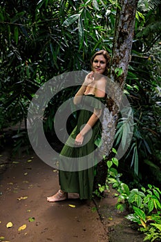 Caucasian woman leaning against a tree in tropical rain forest. Beautiful woman wearing long green dress. Walking trail in jungle