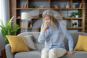 Caucasian woman with low immunity getting seasonal cold and sneezing in paper tissue at home. Mature lady trying to deal