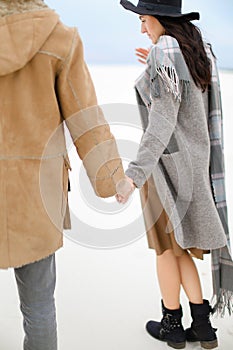 Caucasian woman and man walking on snow and holding hands, wearing coat, grey scarf and hat.